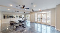 Located in Clayton, NC, this single-floor fitness center is thoughtfully designed to support a full range of workout routines. The space is anchored by striking polished epoxy flooring in a swirling gray marble finish, lending a sleek, modern aesthetic throughout. Multiple ceiling fans are distributed across the room, ensuring consistent airflow and comfort during workouts. Large windows and glass-paneled French doors line the perimeter, flooding the space with natural light and offering views of the surrounding outdoor grounds. Full-length mirrors are mounted along the walls, visually expanding the room and aiding in form correction. The cardio zone features treadmills and cycling bikes positioned near the windows, while a dedicated strength area includes a multi-station cable machine system and a dumbbell rack stocked with a wide range of free weights. Adjustable weight benches are available throughout. A small utility area with cabinetry provides additional convenience. The neutral beige walls and clean white trim complement the overall bright and open atmosphere, making this fitness center a well-rounded and inviting space for daily exercise.