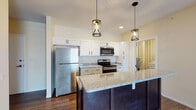 Located at 1200 119th St in Whiting, Indiana, this single-floor residence offers a thoughtfully designed layout with modern finishes throughout. The open-concept kitchen serves as the centerpiece of the space, featuring crisp white shaker cabinetry, granite countertops, and a full suite of stainless steel appliances including a refrigerator, range, and over-the-range microwave. A substantial kitchen island with a dark espresso base and matching granite surface provides additional prep and seating space, while two stylish cage-style pendant lights hang above, adding an industrial-modern touch. Recessed lighting complements the overall brightness of the kitchen area. Rich, dark hardwood-style flooring flows consistently throughout the main living areas, creating a warm and cohesive aesthetic. The open living area is spacious and well-lit, with access to additional rooms and a view into the bathroom. A dedicated bedroom offers a window with natural light and views of the surrounding neighborhood. The bathroom is finished with a white vanity, granite countertop, undermount sink, framed mirror, and warm overhead lighting set against earthy-toned walls. Tile flooring in the bathroom provides a clean contrast to the hardwood found elsewhere, completing this well-appointed, move-in-ready space.