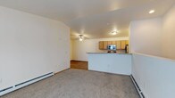 This single-floor residence offers a practical and well-designed open-concept layout that seamlessly connects the living area and kitchen. The living space is finished with plush wall-to-wall carpeting, contributing to a warm and comfortable atmosphere, while baseboard heating units line the walls for efficient climate control. A ceiling fan adds both functionality and a casual aesthetic to the room.

The kitchen is defined by a raised breakfast bar counter with a granite-style surface, providing a natural division between the cooking and living areas while maintaining the open feel. Warm-toned wood cabinetry offers ample storage, and the kitchen is equipped with stainless steel appliances, including a microwave and refrigerator. Flush-mount ceiling lighting illuminates the space effectively.

Adjacent to the main living area, a secondary space features rich hardwood-style flooring, adding a contrasting texture and visual interest to the overall layout. The walls throughout are finished in soft, neutral tones, creating a bright and inviting ambiance. The thoughtful combination of carpet, hard flooring, and functional kitchen design makes this a versatile and comfortable single-level living space suited to a variety of lifestyles.