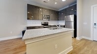 Located at 1000 Central Ave in Charlotte, NC, this one-bedroom, one-bathroom unit offers a thoughtfully designed single-floor layout that makes excellent use of its compact footprint. Light wood-style flooring flows consistently throughout every room, creating a cohesive and inviting aesthetic anchored by neutral, warm-toned walls and clean white baseboards.

The kitchen is a standout feature, showcasing high-gloss gray cabinetry, a classic white subway tile backsplash, and a full suite of stainless steel appliances including a refrigerator, range, and microwave. A central island with granite countertops provides ample prep space and doubles as a casual dining or gathering area, seamlessly connecting the kitchen to the living space.

The living room is bright and open, highlighted by French doors fitted with blinds that invite natural light while offering privacy. A ceiling fan adds both comfort and character to the space.

The bedroom is a comfortable, well-proportioned retreat featuring windows with blinds that allow for adjustable natural light. The room offers a clean, quiet ambiance well-suited for rest.

The bathroom is finished with consistent attention to detail, featuring a dark-toned vanity with granite countertops, a large mirror, and a tiled shower-tub combination. Wood-look tile flooring carries the warm aesthetic of the rest of the home into this functional space.