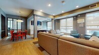 This single-floor club room is a well-appointed communal space designed with a contemporary aesthetic and a range of functional zones suited for social interaction and relaxation. The open layout is unified by warm hardwood-style flooring and a neutral gray-toned color palette accented by bold pops of color throughout.

The main lounge area features a generously sized sectional sofa positioned atop a patterned area rug, complemented by a pair of mid-century-inspired teal patterned accent chairs positioned near large windows fitted with roller shades. These windows allow ample natural light to filter in while providing views of the surrounding neighborhood. A linear fireplace set against a floor-to-ceiling tile surround serves as a refined focal point, with multiple flat-screen televisions mounted nearby for entertainment.

Adjacent to the lounge is a dining area furnished with a sleek rectangular table surrounded by vibrant red chairs, illuminated by a sculptural pendant light. Black-framed glass partitions define this zone while maintaining visual connectivity to the broader space.

A compact kitchenette equipped with dark cabinetry, light countertops, a sink, and under-counter appliances supports event and hospitality needs. Recessed lighting throughout ensures consistent, well-distributed illumination across all areas.