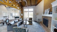 This single-floor space makes a striking first impression with its dramatic vaulted ceiling adorned with exposed wooden trusses and natural wood paneling, creating a warm yet grand architectural character. The open living area is anchored by a full brick fireplace, lending rustic charm alongside the industrial-style exposed ductwork that adds a layer of visual interest overhead.

Large windows rise toward the peak of the ceiling, flooding the interior with generous natural light while offering views of the surrounding greenery outdoors. The bright, neutral walls contrast beautifully with the rich tones of the brick and wood elements throughout.

The seating arrangement is thoughtfully composed, featuring a leather sofa, patterned accent chairs, and a central round coffee table atop a textured area rug, creating an inviting and functional gathering space. The open layout flows naturally between areas, enhancing the sense of spaciousness throughout.

Additional details such as a floor lamp, decorative accessories, and carefully selected furnishings contribute to the polished yet approachable ambiance. The overall design successfully blends rustic, industrial, and contemporary elements, resulting in a space that feels both comfortable and architecturally distinctive.