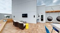 Located at 400 South Greene Street in Greenville, North Carolina, this space showcases a well-appointed rooftop deck alongside a spacious interior corridor. The rooftop deck is a standout feature, finished with warm wood tile decking and framed by clean, modern panel facades. It offers a comfortable outdoor lounge area complete with cushioned wicker seating, a bold yellow accent side table, and a wall-mounted flat-screen television, making it an ideal gathering space for residents or occupants. Built-in wooden bench seating lines one perimeter wall, providing additional capacity for group use — consistent with the posted maximum occupancy signage visible at the stairwell entry. Access to the rooftop is provided via a glass-paneled door connected to a stairwell marked as Stair C, Floor 5. The interior corridor is long and efficiently designed, featuring herringbone-patterned carpet in muted grey and yellow tones that add a subtle visual interest underfoot. Recessed panel ceiling lights illuminate the hallway evenly from end to end, while neutral grey walls and white-trimmed doors maintain a clean, professional aesthetic throughout. Together, these spaces reflect a modern, multi-story building designed with both functionality and communal comfort in mind.