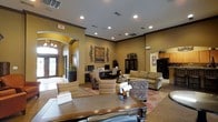 This single-floor community space offers a warm and inviting atmosphere, thoughtfully designed to serve both functional and social purposes. The main area features an open-concept layout with warm golden-toned walls, elegant crown molding, and recessed lighting that creates a bright and welcoming ambiance throughout.

A comfortable lounge area anchors the central space, furnished with plush sofas and accent chairs arranged around a decorative area rug, ideal for relaxed gatherings or informal meetings. Decorative wall art and botanical metal accents contribute to the refined yet approachable aesthetic.

A dedicated business center is clearly visible within the space, equipped with desktop computers and a workstation, offering a convenient resource for residents or visitors. Arched entryways with classic architectural detailing frame the transitions between areas, adding a sense of elegance and depth to the layout.

Near the entrance, a tiled foyer with a chandelier and glass-panel doors allows natural light to filter in, enhancing the open feel of the interior. A kitchenette or refreshment area is also visible toward the rear, complete with appliances and cabinetry, further supporting the multi-functional nature of the space.

Overall, the design balances comfort and practicality, making this a versatile and well-appointed community hub.