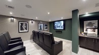 This thoughtfully designed private home theater delivers an immersive cinematic experience within a refined, single-level space. The room is anchored by rows of plush black leather recliner seats, each equipped with integrated cup holders, arranged to face a large wall-mounted flat-screen display. A second large-format screen is mounted on an adjacent deep green accent wall, offering flexible viewing angles throughout the space.

The interior design strikes a bold contrast between the rich forest green accent walls and the textured, neutral grey wallcovering that lines the seating area, creating a warm yet dramatic atmosphere. Recessed ceiling lighting and wall-mounted sconces provide layered, controllable illumination suited for screening events. Light-toned carpet tiles with a subtle linear pattern cover the floors, adding comfort underfoot while enhancing the room's clean, modern aesthetic.

A dedicated snack and beverage station, complete with cabinetry and what appears to be a popcorn machine, adds a genuine theater-going experience to the space. The walls are adorned with a curated collection of large-format black-and-white framed photographs, lending the room an elegant, gallery-like quality. A structural column is seamlessly integrated into the layout, and a ceiling-mounted projector is also present, further enhancing the room's entertainment capabilities. The overall ambiance is polished, inviting, and purpose-built for cinematic enjoyment.