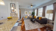 This well-designed single-floor residence offers a cohesive blend of modern style and comfortable living. The open-concept layout connects the kitchen, dining, and living areas seamlessly, creating an inviting and functional flow throughout.

The living room is a standout space, featuring rich hardwood flooring, crown molding, and large windows that allow abundant natural light to pour in. A ceiling fan adds both practicality and character, while a glass-top coffee table and plush area rug anchor the seating arrangement of a charcoal sofa and contemporary accent chairs adorned with bold yellow cushions.

The kitchen is well-equipped with dark cabinetry, granite countertops, stainless steel appliances including a refrigerator, microwave, and range, complemented by a tile backsplash and a breakfast bar with bar seating. Adjacent to the kitchen, the dining area features a glass-top table surrounded by elegant white chairs, bathed in natural light from nearby windows.

The bedrooms are calm and tastefully finished, each with crown molding, hardwood floors, and windows dressed with classic blinds and curtains. The bathroom mirrors the home's refined aesthetic with granite countertops, dark wood cabinetry, and polished chrome fixtures.