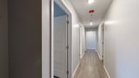 Located at 213 Chenango St in Binghamton, NY, this single-floor property offers a thoughtfully updated interior with a clean, modern aesthetic throughout. A long central corridor with light gray wood-look flooring and a white paneled ceiling connects the various rooms, providing an organized and functional layout.

The kitchen is a standout feature, boasting white shaker-style cabinetry, quartz countertops, stainless steel appliances including a range and refrigerator, and a striking exposed brick accent wall with a professional-grade range hood — adding an industrial character to the space.

Multiple bedrooms are present throughout, each featuring soft carpet flooring, ceiling fans, and natural light from double-hung windows with exterior views. Closets with hanging rods provide practical storage. Living areas display elevated ceilings with decorative beams, light blue-gray walls, wood-look flooring, and generous window arrangements that allow abundant natural light.

The bathrooms are well-appointed, featuring curved glass walk-in shower enclosures, pedestal and vanity sinks, tile flooring, and updated fixtures and lighting. Fire suppression sprinkler systems are visible in the corridor, reflecting attention to building safety standards. Overall, the space presents a clean, functional, and updated environment suitable for multi-occupant use.