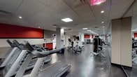 This fitness center is a fully outfitted, single-floor facility designed to meet a wide range of workout needs. The space features a bold and energizing color scheme, with striking red accent walls contrasting against neutral gray tones throughout, creating a motivating atmosphere for exercise.

The cardio zone is prominently positioned along one side of the room, housing a row of Matrix treadmills alongside elliptical trainers and stationary bikes. These machines are arranged to allow for comfortable spacing between users. Toward the back of the facility, a dedicated strength training area offers a selection of cable machines and resistance equipment, providing options for full-body conditioning.

The flooring consists of durable, speckled rubberized material suited for high-traffic fitness use, while the dropped ceiling is fitted with recessed lighting and flush panel lights that ensure the space is evenly and brightly illuminated throughout. Ventilation is well-integrated into the ceiling grid, supporting a comfortable indoor environment during workouts.

The layout is open and thoughtfully organized, allowing for smooth circulation between equipment zones. A mirrored wall at the rear of the strength area enhances the sense of depth and space. Overall, the facility presents a clean, professional, and functional environment well-suited for daily fitness routines.