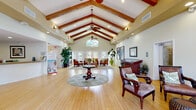 This single-story community clubhouse is defined by its impressive architectural design and warm, resort-inspired ambiance. The centerpiece of the facility is a sweeping great room showcasing soaring vaulted ceilings adorned with rich exposed wooden beams, ceiling fans, and an abundance of recessed lighting. Light hardwood flooring runs throughout, complementing the soft neutral wall tones and lush tropical greenery that accent the space. A central seating area with elegant wooden furniture anchors the room, while large glass doors and a clerestory window at the gable end flood the interior with natural light and offer views of the outdoor surroundings, including a glimpse of a pool area beyond.

Adjacent to the great room, a comfortable lounge area near the entry features refined upholstered chairs and ornate wooden furnishings, creating an inviting gathering spot. A reception desk with a granite countertop and nautical décor accents serves as a welcoming point of contact, accompanied by a display area featuring framed botanical artwork. The fitness center is well-appointed with a comprehensive array of cardio machines, free weights, and strength equipment, mirrored walls to maximize the sense of space, and a ceiling fan to maintain comfort during workouts. Throughout the facility, thoughtful décor, plentiful greenery, and an open layout contribute to a polished and hospitable community environment.