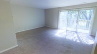 This single-floor space features a generously sized living area finished with neutral-toned wall-to-wall carpeting that lends a clean, understated aesthetic throughout. The walls are painted in a soft, light gray tone, contributing to an open and airy atmosphere. A standout feature of the room is the full-width sliding glass door fitted with vertical blinds, which serves as the primary source of natural light. The door provides direct access to an outdoor area, where surrounding greenery is visible, adding a sense of connection to the outdoors. Sunlight streams through the glass panels, casting bright patterns across the carpet and enhancing the overall sense of spaciousness. The room's layout is straightforward and versatile, offering ample floor space suitable for a variety of furniture arrangements. The smooth, flat ceiling with subtle vent placement keeps the design clean and functional. White baseboards frame the perimeter of the room, providing a crisp contrast to the soft wall color. Overall, the space presents a bright, practical, and well-proportioned environment with a calm, neutral palette.