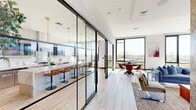 Located at 355 Broad St in Newark, NJ, this single-floor space showcases a sophisticated modern design with generous square footage and an abundance of natural light throughout. The open layout is anchored by floor-to-ceiling black-framed windows that frame sweeping panoramic views of the Newark skyline and surrounding cityscape, flooding the interior with daylight.

The kitchen is a standout centerpiece, enclosed by striking steel-framed glass partition walls that lend an industrial-chic aesthetic while maintaining visual openness. A large marble waterfall island with seating for multiple guests serves as the focal point, complemented by premium stainless steel appliances, a professional-grade range with a stainless hood, built-in undercounter refrigeration, and warm brass pendant lighting overhead.

The living room is equally impressive, featuring a dramatic suspended central fireplace as a bold architectural statement. Corner floor-to-ceiling windows wrap the space in light and offer unobstructed city views. Rich textured accent columns add warmth and visual depth, while a built-in shelving alcove provides both function and character.

Light wood-plank flooring runs throughout, tying the spaces together cohesively. The overall ambiance is one of upscale urban living—refined, open, and thoughtfully designed.