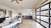 Located at 109 Alicia Way in LaGrange, Georgia, this single-floor open-concept space offers a versatile and thoughtfully designed layout ideal for gathering and entertaining. The interior is defined by polished concrete floors that run throughout, crisp white walls, and recessed lighting that creates a bright, clean aesthetic.

A full-width glass panel garage door serves as a dramatic architectural focal point, flooding the interior with natural light and providing a seamless visual connection to the outdoor deck and surrounding views. Additional windows along the perimeter further enhance the sense of openness and airiness. Ceiling fans are positioned throughout to ensure comfort.

The space is organized into distinct functional zones. A lounge area features a sleek, low-profile linear fireplace flanked by large windows overlooking the outdoors, creating an inviting setting for relaxed seating. A dedicated dining area accommodates a round marble-top table with ample surrounding space. A bar and kitchenette area includes gray cabinetry, a sink, counter seating with bar stools, and a beverage refrigerator, making the space well-equipped for hosting. A side door with glass panels provides convenient access to the exterior. The overall ambiance is modern and refined, with a neutral palette and clean lines throughout.