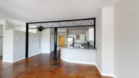 This unique single-floor space blends industrial character with modern comfort through a series of thoughtfully designed areas. Rich, stained concrete floors run throughout, lending warmth and visual depth to every room.

The open living area is an immediate standout, anchored by a striking mid-century style freestanding fireplace with a bold black chimney pipe extending to the ceiling. It rests on a brick hearth pad, adding a rustic contrast to the polished floors. Multiple windows along the exterior wall fill the space with natural light and offer views of surrounding greenery, while a ceiling fan ensures year-round comfort.

A defining architectural feature is the industrial-style steel and glass partition that separates the living area from the kitchen. Its geometric cross-brace design and sliding panel create a dramatic visual boundary while maintaining an open, connected feel.

The kitchen is well-appointed with white shaker cabinetry, gray granite countertops, a subway tile backsplash, and a full suite of stainless steel appliances including a refrigerator, range, microwave, and dishwasher. A curved breakfast bar peninsula provides additional workspace and casual seating.

The bathroom continues the space's cohesive design with granite countertops, white cabinetry, and a tub-shower combination tiled in classic white with a black-and-white checkered accent border. The stained concrete flooring carries through, tying the entire unit together.