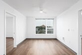 This single-floor unit presents a clean, modern aesthetic defined by crisp white walls, warm hardwood-style flooring, and a well-considered layout throughout. The open living area is a standout space, featuring a large picture window framing elevated views of a hillside cityscape, complemented by a ceiling fan overhead. Natural light flows generously into the room, enhancing its airy, open feel.

The kitchen is efficiently designed in a galley configuration, featuring light natural wood cabinetry, white countertops, stainless steel appliances including a dishwasher, and ample upper and lower storage. The layout maximizes functionality while maintaining a cohesive, minimalist appearance.

Multiple bedrooms are present throughout the unit, each finished with the same warm hardwood-style flooring and neutral white walls. Several bedrooms benefit from windows with outdoor views, while a ceiling fan provides additional comfort in the primary bedroom. A secondary bedroom offers a tall, narrow window admitting natural light.

The bathroom includes a full soaking tub with a clean white surround, brushed nickel hardware, and a simple, understated finish. The entry area provides a welcoming transition into the space, with clear sightlines connecting to the kitchen and living zones. Overall, the unit maintains a consistent, contemporary character throughout.