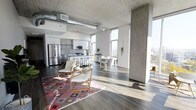 This urban loft showcases a striking industrial-modern aesthetic defined by soaring concrete ceilings with exposed silver ductwork and raw structural columns that lend an authentic, warehouse-inspired character. The single-floor open-concept layout flows effortlessly from the living area through the dining space and into the kitchen, creating a cohesive and airy environment ideal for both everyday living and entertaining. Wide-plank dark hardwood flooring grounds the space and contrasts beautifully with the bright white walls and light-toned finishes throughout. The modern kitchen features stainless steel appliances, sleek white cabinetry, a subway tile backsplash, and a breakfast bar with seating, making it both functional and stylish. The dining area comfortably accommodates a full table and chairs, while the living area is anchored by a vibrant geometric area rug that adds warmth and color. Floor-to-ceiling windows along the perimeter flood the interior with generous natural light and frame panoramic city views, reinforcing the elevated urban setting. The combination of raw architectural elements, contemporary furnishings, and abundant sunlight gives this loft a dynamic and sophisticated ambiance throughout.