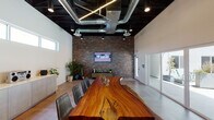 This single-floor commercial space blends industrial and contemporary design elements to create a distinctive and functional meeting environment. The centerpiece of the room is a large live-edge wood conference table, showcasing natural grain and organic contours, surrounded by sleek gray leather chairs. The dramatically dark ceiling features exposed ductwork, linear pendant lighting, and recessed spotlights, creating a bold industrial contrast against the crisp white walls.

A full-height brick accent wall anchors one end of the room, hosting a wall-mounted display screen and flanked by greenery, adding warmth and texture to the space. Opposite, expansive glass sliding doors flood the interior with natural light and provide direct access to an outdoor courtyard lined with planters and seating. A continuous band of clerestory windows along the upper walls further enhances natural light throughout.

A built-in credenza with light wood cabinetry lines one wall, offering functional storage and display space. A separate utility area includes a double-basin sink with brass fixtures, light wood cabinetry, and white tile backsplash. Polished concrete floors tie the space together, reinforcing the clean, modern-industrial aesthetic throughout.
