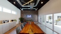 This single-floor commercial space blends industrial and contemporary design elements to create a distinctive and functional meeting environment. The centerpiece of the room is a large live-edge wood conference table, showcasing natural grain and organic contours, surrounded by sleek gray leather chairs. The dramatically dark ceiling features exposed ductwork, linear pendant lighting, and recessed spotlights, creating a bold industrial contrast against the crisp white walls.

A full-height brick accent wall anchors one end of the room, hosting a wall-mounted display screen and flanked by greenery, adding warmth and texture to the space. Opposite, expansive glass sliding doors flood the interior with natural light and provide direct access to an outdoor courtyard lined with planters and seating. A continuous band of clerestory windows along the upper walls further enhances natural light throughout.

A built-in credenza with light wood cabinetry lines one wall, offering functional storage and display space. A separate utility area includes a double-basin sink with brass fixtures, light wood cabinetry, and white tile backsplash. Polished concrete floors tie the space together, reinforcing the clean, modern-industrial aesthetic throughout.
