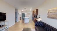 This well-designed single-floor space showcases a thoughtfully laid-out open-concept floor plan that seamlessly connects the living and kitchen areas. The light hardwood flooring runs throughout, creating a warm and cohesive aesthetic that ties the space together.

The kitchen serves as the centerpiece of the home, featuring rich walnut-toned cabinetry, stainless steel appliances, and a white central island with bar seating — ideal for casual dining or entertaining. Pendant lighting hangs above the island, adding a modern touch, while recessed ceiling lights provide ample illumination throughout. A built-in desk or workspace is tucked neatly along one wall, offering a practical and organized area for everyday tasks.

The living area flows directly from the kitchen, furnished with a tufted leather sofa and a area rug, contributing to the comfortable yet polished ambiance. Neutral light gray walls keep the space feeling bright and airy, complementing the natural tones of the flooring and cabinetry.

Additional rooms are accessible via hallways visible from the main living space, including what appears to be a bathroom and further private areas. A distinctive teal-painted interior door adds a charming accent detail. Overall, this space balances modern functionality with a welcoming, homey character.