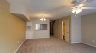 This two-story residence offers a practical and comfortable layout designed for everyday living. The main level centers around a spacious carpeted living area, finished with neutral beige tones that create a warm, welcoming atmosphere. A textured ceiling adds visual interest overhead, while a ceiling fan with integrated lighting promotes year-round comfort. A decorative chandelier serves as a focal point, lending a touch of elegance to the open space. A built-in wall-mounted HVAC unit ensures efficient climate control throughout the main living area. A pass-through window connects the living space to the kitchen, fostering an open, interactive flow between the two areas. Staircase access to the upper floor is conveniently situated within the main living space, providing a seamless transition between levels. A door off the main living area leads to a bathroom, adding practical convenience to the ground floor. The neutral color palette, soft carpeting, and functional design elements come together to create a cohesive and inviting living environment throughout this two-floor home.