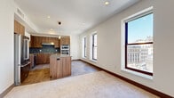 This well-appointed single-floor residence offers three bedrooms and two bathrooms within a thoughtfully designed layout. The open-concept kitchen and living area serves as the heart of the home, featuring warm walnut-toned cabinetry, a tiled backsplash, granite-topped center island, stainless steel appliances including double wall ovens, and pendant lighting. Tile flooring anchors the kitchen zone while plush carpet extends into the living area, creating a natural distinction between spaces. Multiple tall, dark-framed windows line the living area walls, flooding the space with natural light and framing elevated urban views of surrounding city architecture, including a distinctive domed building.

The bedrooms are carpeted and calm in tone, each featuring dark-framed windows with city outlooks and closet access. The primary bathroom is generously proportioned, showcasing large-format stone-look tile flooring, a walk-in glass-enclosed shower with tiled surround, and warm wood vanity cabinetry with a granite countertop. Hallways feature tile flooring and clean white walls, connecting rooms efficiently. Recessed lighting and neutral finishes throughout lend the space a modern, cohesive aesthetic.
