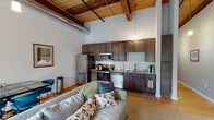 Located at 1151 Falls Rd in Rocky Mount, NC, this single-floor loft-style space showcases a striking blend of industrial character and modern comfort. Dramatic soaring ceilings with exposed wooden beams and visible ductwork define the open living area, which is bathed in natural light streaming through large multi-pane windows fitted with roller shades. Hardwood floors run throughout, anchoring the warm, industrial aesthetic.

The open-concept kitchen is thoughtfully appointed with dark espresso cabinetry extending to ceiling height, granite countertops, a classic white subway tile backsplash, and a full suite of stainless steel appliances including a range, microwave, dishwasher, and refrigerator. A pendant light fixture adds a refined industrial touch overhead.

The spacious living area accommodates generous seating and a dining space, with exposed concrete block walls adding authentic loft character alongside the wood-paneled ceiling details.

The primary bedroom features soaring ceilings, hardwood floors, and direct access to a private en-suite bathroom finished with a marble-topped vanity and a tiled walk-in shower. A secondary bedroom with classic hardwood floors offers dual twin beds with decorative headboards. The clean, well-appointed bathroom rounds out this thoughtfully designed urban loft retreat.
