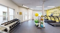This fitness center at 2225 Ashley River Rd in Charleston, South Carolina, offers a comprehensive workout facility designed to support a wide range of exercise routines. The single-floor layout is thoughtfully organized, maximizing the available floor space with a diverse selection of equipment.

The cardio zone features a row of TRUE-brand treadmills, elliptical trainers, and upright stationary bikes, all positioned in front of large windows that allow generous natural light and views of the surrounding greenery. A bold, yellow motivational mural covers the accent wall, displaying a series of encouraging phrases that set an energizing tone throughout the space.

The strength and functional training area includes a multi-station cable machine, an adjustable weight bench, a full rack of dumbbells, and an assortment of medicine balls and stability balls on a dedicated storage stand. Large wall mirrors in this section enhance the sense of openness and allow users to monitor their form during exercises.

The flooring throughout consists of durable rubber gym matting, providing both safety and comfort underfoot. The space is well-lit with recessed and flush-panel ceiling lighting, and a set of French doors at the entry area offer easy access while contributing to the bright, airy atmosphere. The overall design is clean, functional, and motivating — well-suited for daily fitness use.