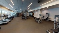 Located at 8051 33rd Ave S in Bloomington, Minnesota, this well-appointed fitness center delivers a comprehensive and contemporary workout environment. The space is thoughtfully designed with a clean, modern aesthetic, featuring a drop ceiling with linear LED lighting and ceiling fans that maintain a bright, comfortable atmosphere throughout.

The main workout floor is generously sized, offering ample room for a wide array of Life Fitness strength machines, cable systems, free weight stations, and a punching bag. A dedicated cardio zone houses multiple treadmills and a stair climber, all positioned alongside large windows that bring in natural light and provide views of the surrounding outdoor area.

At the entrance, a well-organized service station features built-in shelving with under-counter storage, a mini refrigerator, and neatly arranged cleaning supplies, ensuring the space remains sanitary and welcoming. Fitness center rules are posted prominently near the door for easy reference.

Adjacent to the workout floor, a glass-walled lounge area furnished with seating and pendant lighting provides a space to relax before or after exercise. The transition between zones is seamless, with floor-to-ceiling glass partitions maintaining visual connectivity while defining each area. Together, these elements create a professional, fully equipped fitness facility suited for a wide range of training needs.