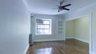 This single-floor space showcases a blend of classic architectural character and functional design. The main living area features rich hardwood floors that reflect natural light streaming in through a large, white-trimmed window fitted with a window air conditioning unit. Crown molding lines the ceiling, adding an elegant, prewar-style touch to the room's overall aesthetic.

A ceiling fan with a brushed metal finish is centrally mounted, providing both airflow and a subtle modern contrast to the room's traditional details. Built-in open shelving is recessed into one wall, offering dedicated display or storage space while maintaining a clean, streamlined look. A classic cast-iron radiator beneath the window speaks to the building's heritage and serves as a functional heating element.

The walls are painted in a soft, neutral gray with a partial accent of warm sage green in an adjoining area, creating a sense of visual separation between spaces. Crisp white baseboards and door trim throughout reinforce the traditional detailing. Overall, the space offers a warm, understated ambiance that balances historic architectural charm with everyday livability.