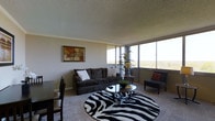 This single-floor residence offers a well-appointed living space filled with natural light, thanks to a striking wall of floor-to-ceiling windows that frame expansive views of the surrounding landscape. The living area is generously sized, centered around a plush dark sofa accented with decorative pillows and paired with a sleek glass coffee table resting atop a bold zebra-print area rug — a design choice that adds a distinct visual character to the room. A comfortable accent chair and a tall indoor plant complete the seating arrangement, contributing to the inviting ambiance. Crown molding and a textured ceiling add classic architectural detail throughout. An adjacent dining area is visible, providing a natural transition between living and entertaining spaces. The unit's elevated position is one of its most notable features — the balcony offers a broad, open-air panorama stretching across tree-lined neighborhoods, parking areas, and commercial buildings under wide, clear skies. This elevated vantage point brings in abundant daylight and a sense of openness that defines the overall feel of the space. The neutral carpet and light-toned walls provide a versatile backdrop suitable for a variety of interior styles.