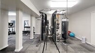 This state-of-the-art fitness center, spanning 930 square feet, is a comprehensive exercise facility designed to cater to various workout needs. The space is thoughtfully divided into distinct zones, each dedicated to specific types of exercise. Upon entry, visitors are greeted by an array of cardio equipment, including treadmills, ellipticals, and stationary bikes, positioned to maximize space efficiency.

The strength training area features a multi-functional cable machine, allowing for a wide range of exercises. Free weights, including dumbbells and barbells, are neatly organized along the walls. A separate section houses weight machines targeting different muscle groups. The center also includes a functional training area with equipment such as stability balls and resistance bands.

The facility boasts modern aesthetics with a neutral color palette, predominantly featuring whites and grays. The flooring consists of durable, low-maintenance carpet tiles in a complementary gray tone. Bright, recessed lighting fixtures ensure optimal visibility throughout the space. Large windows flood the area with natural light and offer views of the outdoors, creating an open and inviting atmosphere.

Wall-mounted televisions and informational signage provide entertainment and guidance for users. The layout promotes easy navigation and maintains clear pathways between equipment. Overall, this fitness center combines functionality with contemporary design, offering a comprehensive workout experience within a compact, well-organized space.