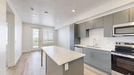 This well-designed one-bedroom, one-bathroom unit in Salt Lake City, Utah offers a thoughtful layout across a single floor. The open-concept kitchen serves as the heart of the space, featuring sleek gray flat-front cabinetry, a white countertop island with additional storage, and a full suite of stainless steel appliances including a refrigerator with ice maker, electric range, over-the-range microwave, and dishwasher. Light wood-style flooring runs throughout the main living areas, enhancing the bright and modern aesthetic. Recessed lighting and white walls keep the space feeling clean and airy. A private balcony accessible from the main living area provides an outdoor retreat with natural light filtering into the interior through large windows fitted with blinds. The bedroom is a comfortable size, finished with plush neutral carpeting and a broad window that invites natural light. Three closets offer generous storage throughout the unit, including a well-organized walk-in closet with multi-level hanging rods and shelving. The bathroom features a soaking tub, wood-style flooring, and a navy vanity. A hallway and utility room round out the functional layout of this contemporary Salt Lake City residence.