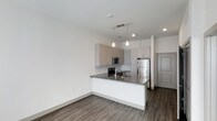 This single-floor residence in The Colony showcases a clean, modern aesthetic with an open-concept layout that seamlessly connects the kitchen and living areas. The kitchen serves as the centerpiece of the space, featuring flat-front cabinetry in a warm taupe finish paired with a classic subway tile backsplash. Dark granite countertops extend across the peninsula, which provides additional prep space and casual seating. Stainless steel appliances, including a refrigerator, built-in microwave, and range, complement the contemporary design. Pendant lighting hangs above the peninsula, adding focused illumination and a stylish accent. Recessed lighting throughout ensures the space remains bright and well-lit. The flooring consists of durable wood-look planks in a medium grey-brown tone, running consistently through the kitchen and living areas for a cohesive look. Neutral white walls keep the space feeling open and versatile. A solid entry door with a modern panel design adds a refined touch at the entryway. The overall ambiance is sleek and functional, reflecting a well-considered contemporary design suited for everyday living.