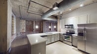This single-floor space showcases a striking industrial aesthetic rooted in its original architectural character. Throughout the main living area, original exposed brick walls with bricked-in window openings are framed by raw concrete columns and beams, creating a rugged, warehouse-inspired atmosphere. The polished concrete floors extend across the expansive open floor plan, complementing the bare concrete ceiling overhead, which features prominently exposed HVAC ductwork, conduit piping, and a ceiling fan that reinforce the industrial character of the space.

The kitchen is thoughtfully appointed with gray shaker-style cabinetry, white quartz countertops, and a classic white subway tile backsplash. Stainless steel appliances include an electric range, over-the-range microwave, and a French door refrigerator. A generously sized peninsula provides additional prep and seating space, while pendant lights suspended from the concrete ceiling add focused illumination. The kitchen opens directly to the broad main living area, enhancing the open-concept flow.

The bathroom features a clean, minimalist design with a white quartz vanity top, undermount sink, brushed nickel faucet, a large mirror, and stone-toned flooring consistent with the rest of the unit. Overall, the space balances raw industrial elements with modern finishes.