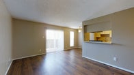 This single-floor residence offers a clean, functional layout with a welcoming open-concept living and dining area. Rich, dark hardwood flooring runs throughout the main living space, adding warmth and a sense of continuity. The living area is bathed in natural light, courtesy of a large sliding glass door fitted with vertical blinds and an adjacent side window, both of which provide views to the outdoors. A ceiling fan with a light fixture is centrally positioned, offering both comfort and supplemental lighting. The textured popcorn ceiling adds a classic residential character to the space. A partial wall with a pass-through counter connects the living area to the kitchen, creating an open flow while maintaining a defined separation between spaces. The kitchen features warm yellow-toned upper cabinetry, giving it a bright and inviting feel. The neutral, tan-toned walls throughout complement both the flooring and the cabinetry, resulting in a cohesive and understated color palette. The overall ambiance is practical and comfortable, with an emphasis on natural light and open sightlines that make the space feel airy and well-connected.