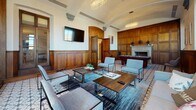 Located at 400 Walnut St in Philadelphia's historic Old City neighborhood, this sophisticated single-floor lounge space exudes classic architectural character blended with contemporary furnishings. The room is defined by floor-to-ceiling dark walnut wood paneling that lines the walls, lending a warm, club-like ambiance throughout. A stunning barrel-vaulted ceiling with ornate plaster detailing draws the eye upward, while elegant oval crystal chandeliers provide refined illumination. The patterned encaustic tile flooring adds a distinctive decorative element that anchors the space with artisanal charm. Tall wood-framed windows and frosted glass panel doors flood the interior with natural light, creating an inviting atmosphere. The lounge is thoughtfully arranged with a generous seating area featuring a plush neutral sofa, sleek modern accent chairs, and wood-topped coffee tables set atop a layered area rug. A wall-mounted flat-screen television serves as a focal point on one end, while a classic marble fireplace and a full-size billiards table offer additional entertainment options. Arched wood doorways with oversized double doors connect the space to adjacent corridors, reinforcing the grand, historic character of this beautifully appointed communal retreat.