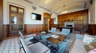 Located at 400 Walnut St in Philadelphia's historic Old City neighborhood, this sophisticated single-floor lounge space exudes classic architectural character blended with contemporary furnishings. The room is defined by floor-to-ceiling dark walnut wood paneling that lines the walls, lending a warm, club-like ambiance throughout. A stunning barrel-vaulted ceiling with ornate plaster detailing draws the eye upward, while elegant oval crystal chandeliers provide refined illumination. The patterned encaustic tile flooring adds a distinctive decorative element that anchors the space with artisanal charm. Tall wood-framed windows and frosted glass panel doors flood the interior with natural light, creating an inviting atmosphere. The lounge is thoughtfully arranged with a generous seating area featuring a plush neutral sofa, sleek modern accent chairs, and wood-topped coffee tables set atop a layered area rug. A wall-mounted flat-screen television serves as a focal point on one end, while a classic marble fireplace and a full-size billiards table offer additional entertainment options. Arched wood doorways with oversized double doors connect the space to adjacent corridors, reinforcing the grand, historic character of this beautifully appointed communal retreat.