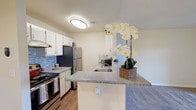 This single-floor space centers around a well-appointed kitchen designed for both practicality and style. The kitchen features crisp white cabinetry that provides ample storage, complemented by a striking blue mosaic tile backsplash that adds a subtle decorative accent. Stainless steel appliances, including a refrigerator and a range with matching hood vent, lend a modern, cohesive look to the cooking area.

The peninsula countertop, finished in a speckled granite-style surface, extends the workspace while creating a natural partition between the kitchen and the adjoining living area. An integrated sink is conveniently positioned within the peninsula, enhancing the kitchen's functionality. The warm-toned wood-look flooring runs throughout, contributing to an inviting atmosphere.

Overhead, a flush-mount ceiling light provides consistent illumination across the space. The textured ceiling and clean white walls keep the overall aesthetic light and neutral. The open layout promotes easy movement between areas, making the space feel connected and efficient for everyday living.