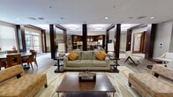 Located at 1901 South Charles Street in Baltimore, MD, this sophisticated single-floor amenity space offers a generously sized, open-concept layout designed for both socializing and relaxation. The space is anchored by a stylish central lounge featuring rich hardwood-style flooring, bold dark columns, and recessed circular ceiling fixtures that cast a warm, inviting glow throughout. The main seating area is furnished with a plush sofa accented by orange throw pillows, coordinating lounge chairs, and a patterned area rug, all arranged around a substantial wood coffee table. A fully equipped kitchen lines the rear of the space, showcasing dark espresso cabinetry, a mosaic tile backsplash, light quartz countertops, stainless steel appliances including a full-size refrigerator, and a central island with pendant lighting. A dedicated dining area features a round wood-grain table with seating for multiple guests and direct access to an outdoor terrace through sliding glass doors. Additional lounge zones offer flat-screen TVs, accent artwork, and intimate seating clusters. An entry foyer with ornate wood doors, clean sightlines, and neutral tones completes this well-appointed communal retreat.