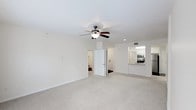 This single-floor residence offers a thoughtfully arranged layout designed for comfortable everyday living. The main living area is generously sized, featuring soft wall-to-wall carpeting in a neutral beige tone that enhances the warmth of the space. A ceiling fan with an integrated light fixture is centrally mounted, providing both ambient lighting and air circulation. The walls are finished in a clean, light cream tone, contributing to a bright and airy atmosphere throughout.

Recessed lighting in the ceiling adds a modern touch, supplementing the natural brightness of the space. An open pass-through connects the living area to the kitchen, which is equipped with stainless steel appliances, including a microwave and oven range, as well as a refrigerator. The kitchen cabinetry is finished in white, and the countertops are visible from the living space, making the layout feel cohesive and functional.

A doorway leads from the main living area to an additional room, likely a bedroom or hallway, offering a clear and practical flow between spaces. The overall design is clean, neutral, and move-in ready.