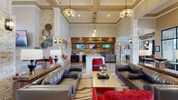 Located in Nashville, TN, this single-story community clubhouse offers a sophisticated and welcoming environment designed for both relaxation and socializing. The expansive main lounge commands attention with soaring coffered ceilings, stacked stone columns, and polished concrete floors that reflect abundant natural light streaming through floor-to-ceiling windows overlooking an outdoor terrace. Seating arrangements throughout include plush leather sofas, vibrant red accent chairs, and patterned area rugs that anchor each gathering space with warmth and style. A standout coffee bar area features a reclaimed wood accent wall, wall-mounted flat-screen TVs, a white quartz countertop with bar seating, and a built-in beverage refrigerator — ideal for casual meetings or everyday refreshment. A secondary lounge room offers a more intimate setting, with globe pendant lighting, eclectic wooden stump stools, bold abstract artwork, and a large mounted television framed by a coffered wood ceiling inset. A separate seating nook near the fireplace showcases a mosaic-tiled surround, elegant wingback chairs, and tall transom windows that fill the space with natural light. Throughout the clubhouse, curated artwork and layered lighting — from drum chandeliers to pendant fixtures — create a cohesive, upscale ambiance.