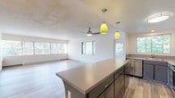This single-floor residence is designed with an open-concept layout that seamlessly connects the kitchen, dining, and living areas. The kitchen is well-appointed with two-tone cabinetry — white upper cabinets paired with grey lower cabinets — complemented by neutral countertops and a full suite of stainless steel appliances, including a refrigerator, electric range, and dishwasher. A central island with a grey base and matching countertop provides additional prep space and casual seating, accented overhead by distinctive yellow pendant lights. A ceiling fan is centrally positioned in the open living area, which benefits from an abundance of natural light through large windows overlooking lush greenery. Light wood-look flooring flows throughout the main living spaces, while built-in shelving adds functional storage along one wall. The bedroom features neutral carpet, a generously sized closet, and a window framing outdoor views. The bathroom includes wood-look flooring, a soaking tub with tiled surround, a white vanity with storage, and a mirrored medicine cabinet. Baseboard heating is present throughout, and a textured ceiling finish is consistent across the space.
