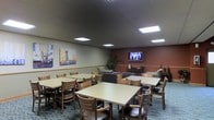 This single-floor communal space functions as a combined dining and lounge area, designed to accommodate group gatherings in a comfortable, accessible setting. The room features multiple rectangular dining tables paired with sturdy wooden chairs with upholstered seats, arranged to maximize seating capacity while maintaining an open, navigable layout. Overhead, a drop ceiling with recessed fluorescent panel lighting provides consistent, even illumination throughout the space.

The room is divided into two distinct zones. The dining area occupies the larger portion, offering ample seating for residents or guests across several table groupings. Toward the back, a cozy lounge area features upholstered seating arranged around a wall-mounted flat-screen television, set against a warm terracotta-colored accent wall that adds visual contrast to the otherwise neutral palette. A wooden media console beneath the TV provides storage and display space.

Colorful abstract artwork adorns the lighter walls, contributing a lively, welcoming character to the room. Indoor plants near the lounge area introduce a touch of greenery. The carpeted flooring throughout adds warmth and helps define the space as a comfortable, community-oriented gathering area.