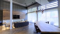 This single-level space showcases a refined contemporary design that seamlessly blends industrial and organic elements. The open-plan layout connects a living area and dining space, creating a cohesive, flowing environment ideal for both relaxation and entertaining.

The living area is anchored by a striking feature wall clad in warm wood paneling, contrasted by a dark square-tiled accent wall that adds texture and depth. A wall-mounted television sits flush against the wood paneling, while a low-profile sofa and area rug define the seating zone. Herringbone-patterned hardwood flooring runs throughout, adding a sophisticated visual rhythm underfoot.

The dining area is highlighted by a substantial wooden table paired with dark chairs, illuminated by a sculptural multi-globe pendant light fixture that serves as a standout design element. Large floor-to-ceiling sheer-draped windows and elevated clerestory windows flood the space with abundant natural light while framing lush outdoor greenery. The combination of high ceilings, dark ceiling detailing, and recessed and track lighting contributes to an atmosphere that is both dramatic and inviting. Overall, the space exudes a modern, upscale ambiance with careful attention to material contrast and architectural detail.