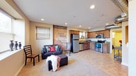 This single-floor space offers an open-concept layout that seamlessly connects the living area and kitchen, creating a functional and inviting environment. The living area is carpeted and comfortably arranged with seating, accented by a bold exposed brick and stone column that serves as a striking architectural focal point, adding character and texture to the space. Recessed lighting throughout the ceiling provides consistent, well-distributed illumination, while a window with horizontal blinds allows for natural light and ventilation. The kitchen flows directly from the living area and features rich wood cabinetry, stainless steel appliances including a refrigerator, electric range, and over-the-range microwave, as well as a sink with a window view above it. The countertops and cabinetry offer ample workspace and storage. The overall design balances a warm, homey atmosphere with practical utility, making the space well-suited for everyday living. The combination of carpeted flooring in the living zone and harder flooring in the kitchen area helps define each functional space within the open plan.