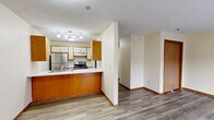 Located at 11581 Raven St NW in Coon Rapids, Minnesota, this single-floor space offers a practical and functional layout comprising a living room, kitchen, hallway, and bathroom. The living room is an open, light-filled area featuring light-toned laminate wood flooring, a baseboard heating unit, a textured ceiling, and a set of wood-framed sliding glass doors that invite natural light and provide outdoor access. The kitchen is designed around a warm wood-paneled peninsula with a built-in sink and white countertop, offering a natural division from the living area. It is equipped with white upper cabinetry, wood-accented lower cabinets, a stainless steel refrigerator, and an electric range. A track lighting fixture overhead ensures the cooking area is well lit. The hallway connects the main living spaces and features additional cabinetry, maintaining storage accessibility throughout. The bathroom includes a combined tub and shower with a curtain rod, a single-sink vanity with wood cabinetry and a large mirror, and overhead lighting. Tile flooring and neutral wall tones give the bathroom a clean, utilitarian character. Textured ceilings are consistent throughout the space, and light-toned laminate flooring flows through the main living areas, contributing to a cohesive and understated aesthetic.