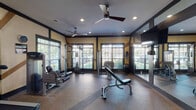 This fitness center is thoughtfully designed to accommodate a full range of workout needs within a single-level layout. The interior draws on a craftsman-inspired aesthetic, featuring bold dark-trimmed structural elements and diagonal accent beams set against warm yellow walls, creating a distinctive and energetic atmosphere. The durable, speckled rubber flooring provides both safety and practicality throughout the space.

The facility is generously equipped with cardio machines, including multiple treadmills and ellipticals by Precor, as well as stationary bikes and a stair climber. Strength training is well represented with a cable machine, adjustable weight bench, dumbbell rack, and additional resistance equipment. Full-length mirrored walls line multiple sections of the room, visually expanding the space and allowing users to monitor their form.

Natural light enters through numerous large windows fitted with horizontal blinds, offering views of lush outdoor greenery. Ceiling fans are installed throughout, supplementing the recessed lighting overhead and maintaining comfortable airflow during workouts. Elegant French doors provide access points that further contribute to the space's refined character, balancing function with polished design.