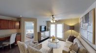 This single-floor residence offers a 1-bedroom, 1-bathroom layout designed for comfortable everyday living. The open living area serves as the heart of the home, featuring a ceiling fan with integrated lighting, warm neutral-toned walls, and a combination of hardwood-style and carpeted flooring that adds texture and warmth underfoot. French doors with slatted blinds allow natural light to fill the space while providing access to the outdoors. The living room comfortably accommodates a full seating arrangement, centered around a round glass coffee table and anchored by a plush area rug, creating an inviting atmosphere for relaxing or entertaining. A media console with a mounted flat-screen television anchors the main wall, complemented by stylish accent lighting. The bedroom, visible through an open doorway, features a generously sized bed with an upholstered headboard and continued neutral tones, maintaining a cohesive and calming aesthetic throughout. The overall ambiance of the space is warm and livable, with a well-balanced blend of natural light, modern furnishings, and neutral finishes that create a welcoming environment suitable for a variety of lifestyles.