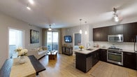 This single-level residence showcases a thoughtfully designed open-concept layout that seamlessly integrates the kitchen, dining, and living areas into one cohesive space. The kitchen is appointed with rich dark cabinetry, light quartz-style countertops, a stainless steel sink, and a dishwasher. A peninsula with bar seating extends into the living area, complemented by elegant pendant lighting overhead. The backsplash adds a clean, modern contrast to the dark cabinetry. Light hardwood-style flooring runs throughout, contributing to an airy, warm ambiance. The living area is generously proportioned, offering space for comfortable seating alongside a rustic wood coffee table and a live-edge bench. Recessed lighting and a ceiling fan ensure both functionality and comfort. Sliding glass doors lead to a private balcony, filling the interior with natural light and offering views of the surrounding area. A bedroom is visible through a connecting doorway, adding a sense of depth to the floor plan. The overall aesthetic balances contemporary design with warm, inviting touches throughout.