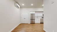 This thoughtfully designed single-floor unit showcases a clean, contemporary aesthetic throughout. The open-concept living and kitchen area is anchored by warm hardwood-style flooring, crisp white walls, and recessed lighting that creates a bright and airy atmosphere. A wall-mounted mini-split system provides efficient climate control directly in the living space.

The kitchen is a standout feature, outfitted with white shaker-style cabinetry accented by gold hardware, light quartz countertops, a white subway tile backsplash, and a full suite of stainless steel appliances — including a refrigerator, electric range, over-the-range microwave, and dishwasher. A stainless steel undermount sink with a modern pull-down faucet completes the functional yet stylish setup. A set of double doors off the living area provides additional storage.

The bedroom is a calm, neutral retreat with the same hardwood-style flooring, soft natural light from a window, and clean white walls. The full bathroom features white penny tile flooring, a tiled walk-in shower, a wall-mounted vanity with a white countertop, and a baseboard heater. The overall design is cohesive and move-in ready, reflecting modern finishes throughout.