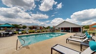 Located at 600 Firestone Ave in Muscle Shoals, Alabama, this single-level outdoor amenity area is designed to serve as a central gathering and recreation hub. At its heart is a generously sized rectangular swimming pool with clear, blue-green water, surrounded by a spacious concrete pool deck lined with multiple lounging chairs and side tables — ideal for relaxing under the Alabama sun.

Adjacent to the pool is a well-appointed covered pavilion featuring a vaulted ceiling with ceiling fans for added comfort. The pavilion is furnished with teal cushioned sectional sofas and accent chairs, creating an inviting outdoor lounge atmosphere. Picnic tables provide additional seating options for casual dining or social gatherings.

The facility also includes a dedicated brick restroom building accessible directly from the covered pavilion area, offering convenience for pool users. Iron perimeter fencing encloses the pool area, providing both safety and a clean aesthetic boundary. Lush green landscaping and mature trees are visible throughout the surrounding grounds, enhancing the open, airy feel of the space. The overall design balances functionality and comfort, making it a well-equipped community recreational destination.